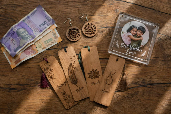 Overhead flat-lay of personalized wooden bookmarks with names, mandala wooden earrings, and an acrylic Best Brother Ever photo frame arranged on a wooden table beside Indian rupee notes