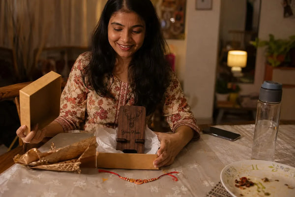 Sister unwrapping a personalized wooden gift beside a rakhi thread on a home dining table the evening before the festival