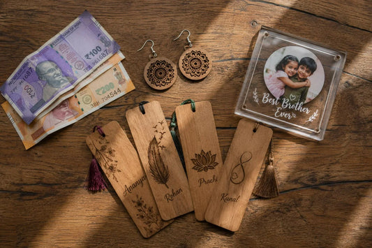 Overhead flat-lay of personalized wooden bookmarks with names, mandala wooden earrings, and an acrylic Best Brother Ever photo frame arranged on a wooden table beside Indian rupee notes