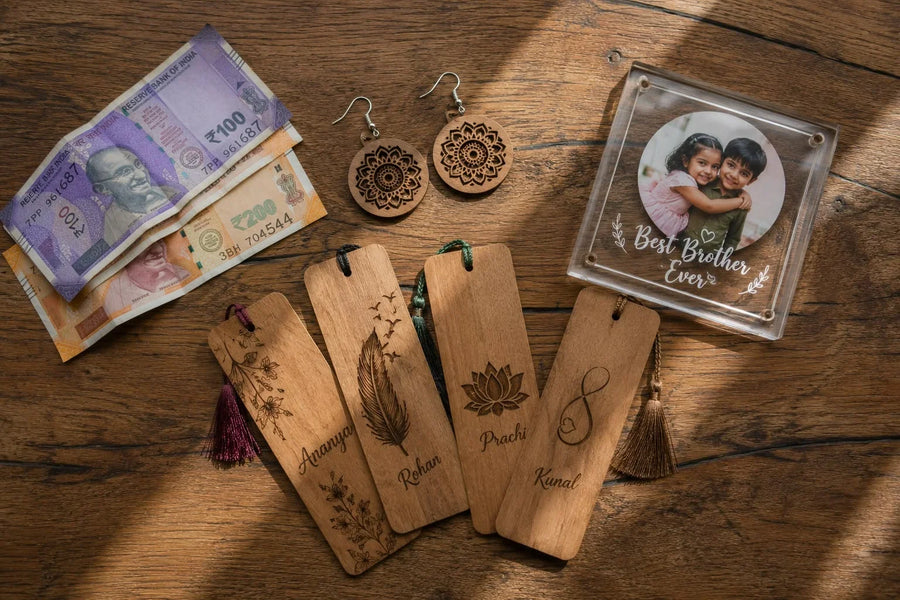 Overhead flat-lay of personalized wooden bookmarks with names, mandala wooden earrings, and an acrylic Best Brother Ever photo frame arranged on a wooden table beside Indian rupee notes