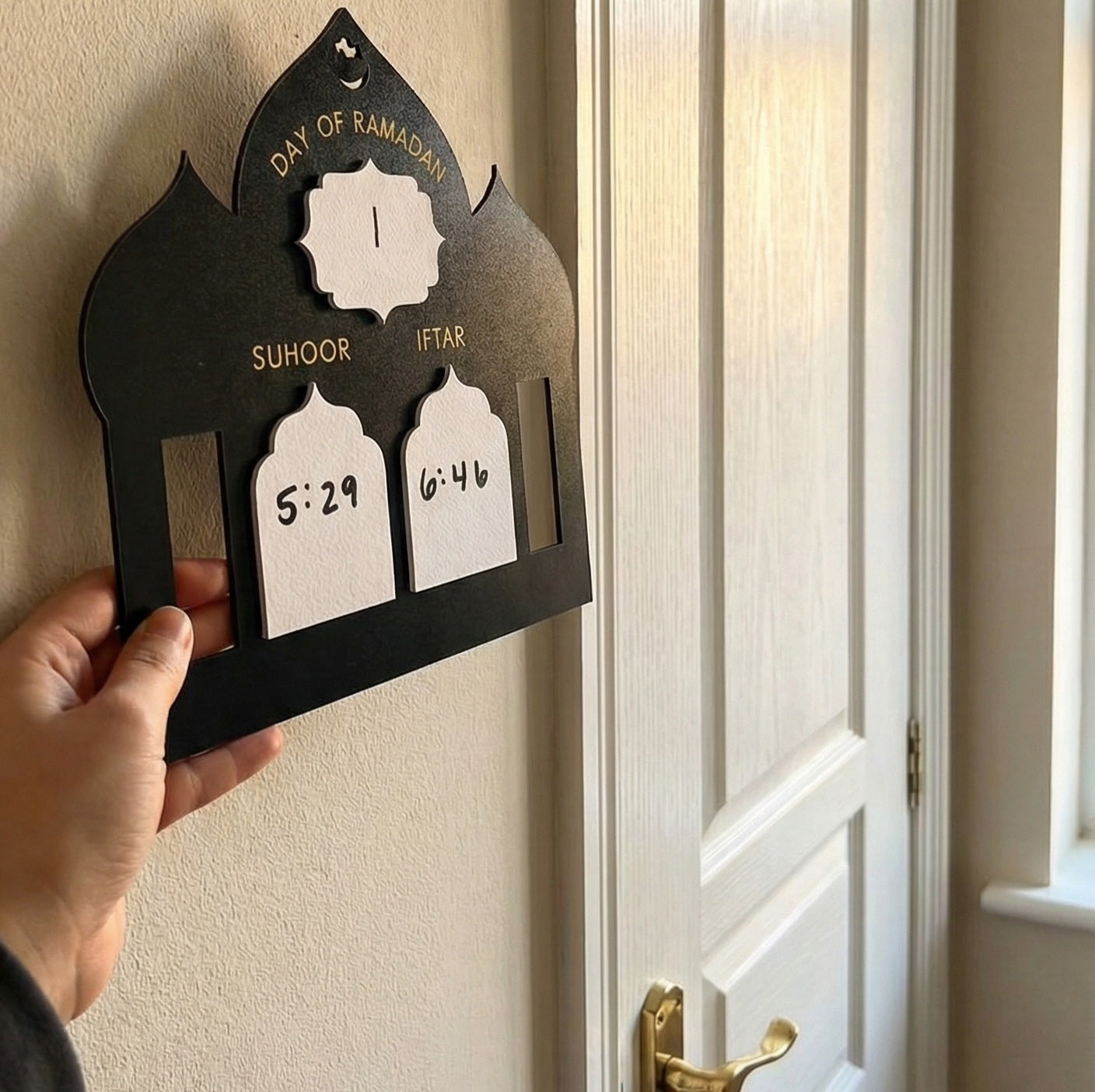 Hand holding a black Ramadan prayer time board against a door.